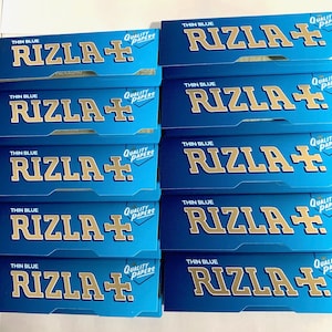 Puede incluir: Una pila de diez papeles de fumar Rizla+ azules y dorados, cada uno con el texto "Thin Blue" y "Quality Papers" impreso en ellos.