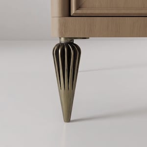 Può includere: Primo piano di una gamba di mobile con un design decorativo scanalato. La gamba è di un colore oro tenue e si assottiglia fino a un punto. Il mobile sembra essere di colore marrone chiaro.