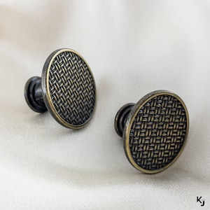 Op de afbeelding: Twee antieke goudkleurige kastknoppen met een geweven patroon. De knoppen zijn rond en hebben een reliëfdesign.