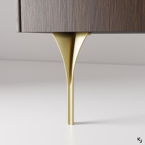 Op de afbeelding: Een close-up van een gouden metalen kastpoot met een uniek gebogen ontwerp. De poot is bevestigd aan een donkerhouten kast.