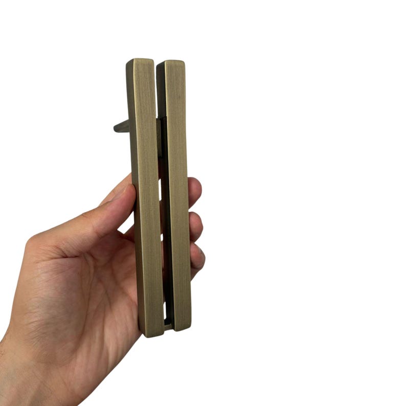 May include: A rectangular, antique brass-colored cabinet pull with a vertical design. The pull is held in a hand against a white background. The pull has a small, protruding handle.