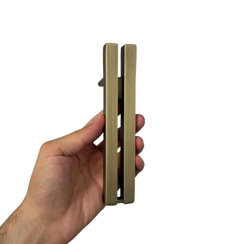 May include: A bronze-colored door handle with a rectangular design. The handle is held vertically against a white background. The handle has a lever and a rectangular shape.