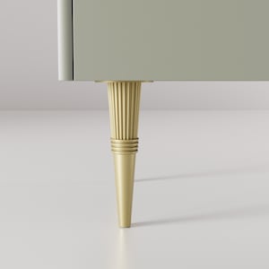 Op de afbeelding: Een close-up van een gouden meubelpoot met een geribbeld ontwerp. De poot is bevestigd aan een lichtgrijze kast.