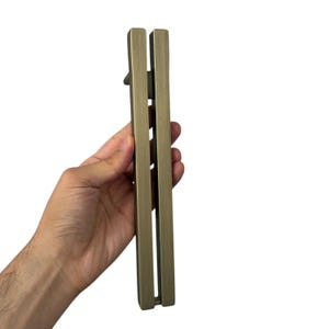 May include: A bronze-colored door pull handle with a rectangular design. The handle is held vertically against a white background. The handle has a sleek, modern design, with a long, narrow shape and a smooth finish. The handle is made of metal.