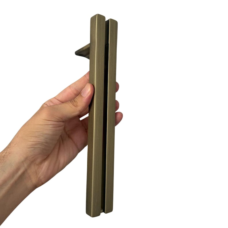May include: A bronze-colored door pull with a rectangular design. The handle is held vertically against a white background. The pull has a simple, modern aesthetic, suitable for various interior design styles.