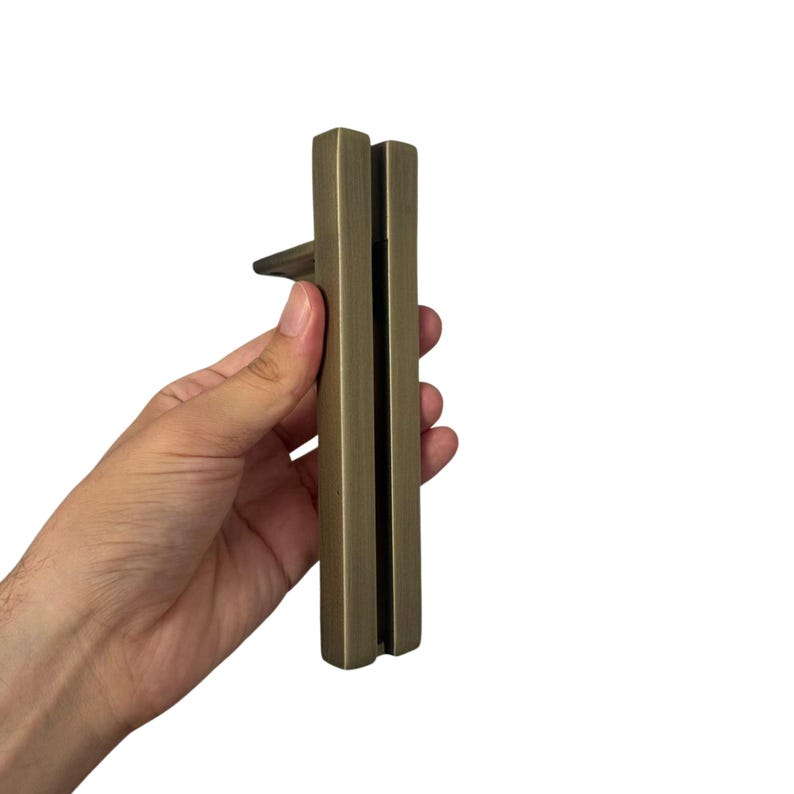 May include: A bronze-colored cabinet pull with a rectangular design. The pull features a small, rectangular handle extending from the main body. The hardware is held in a hand against a white background.