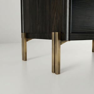 May include: Close-up of a dark wood cabinet with brass-colored metal legs. The cabinet has a rectangular shape with vertical wood grain. The legs are rectangular and have a vertical design. The cabinet is on a white surface.