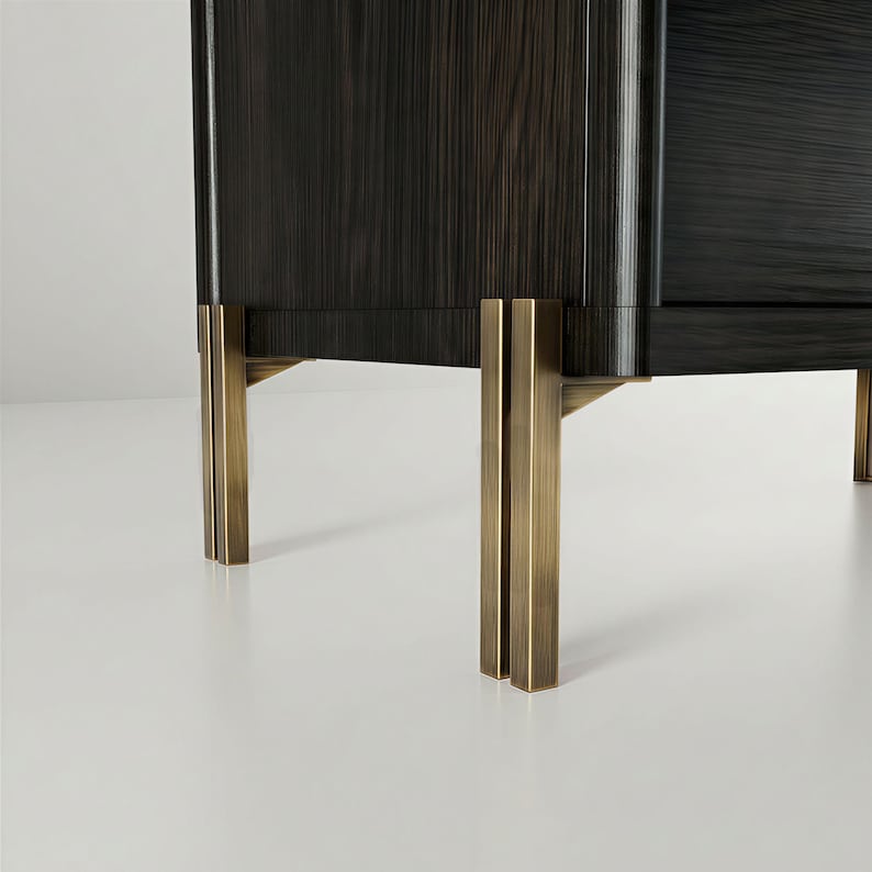 May include: Close-up of a dark wood cabinet with brass-colored metal legs. The cabinet has a rectangular shape with vertical wood grain. The legs are rectangular and have a vertical design. The cabinet is on a white surface.