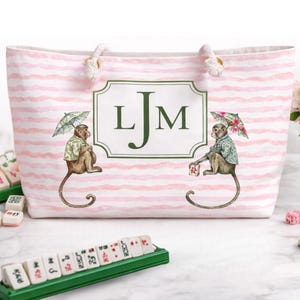 Puede incluir: Bolso tote a rayas rosas y blancas con asas de cuerda. La bolsa presenta un monograma enmarcado en verde y dos ilustraciones de monos sosteniendo paraguas. Las fichas de mahjong son visibles en primer plano.