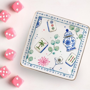 May include: A square white ceramic trinket tray with a gold-trimmed edge, featuring a blue and white ginger jar, mahjong tiles, a martini glass, green eucalyptus sprigs, and a pink flower illustration, surrounded by six translucent pink dice.