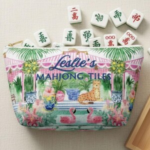 A fabric Mahjong tile bag on a beige background with Mahjong tiles spilling out of the open zipper. The print shows pink flamingos, palms, and blue vases and a custom name