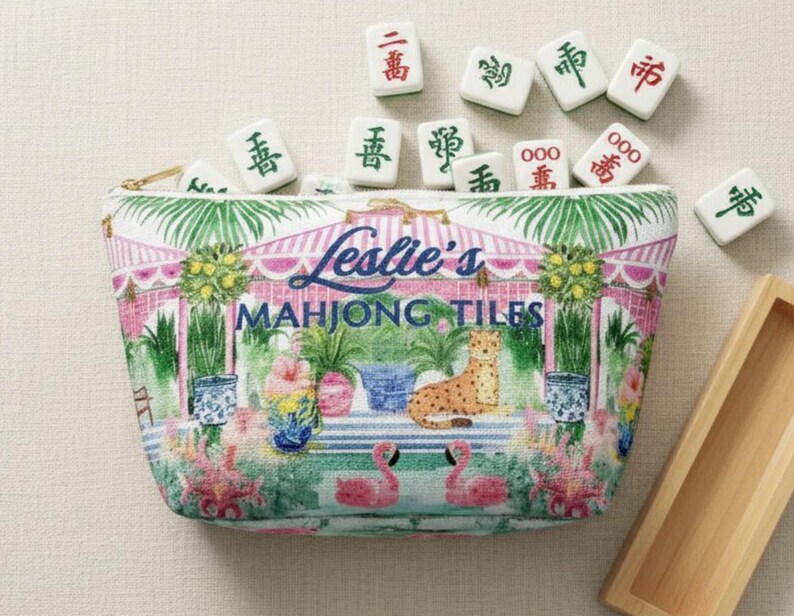 A fabric Mahjong tile bag on a beige background with Mahjong tiles spilling out of the open zipper. The print shows pink flamingos, palms, and blue vases and a custom name