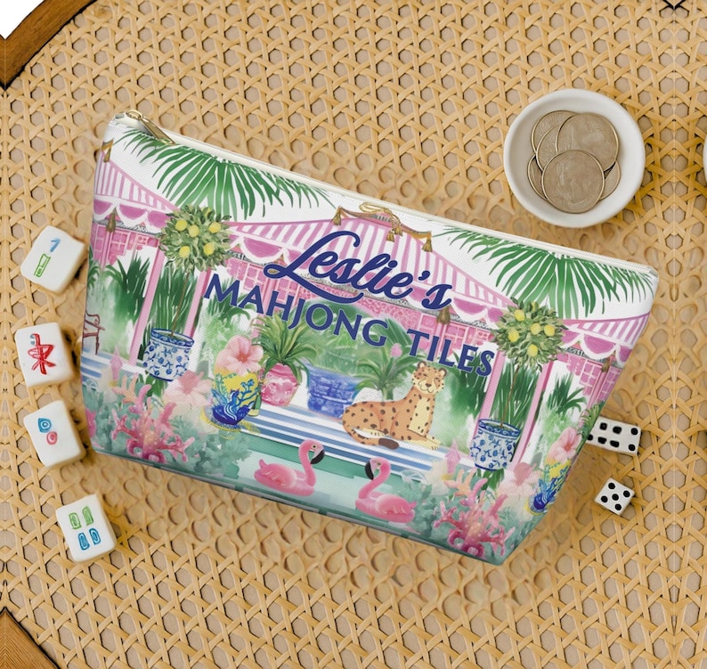 An overhead view of a personalized chinoiserie-design Mahjong tile storage bag surrounded by dice, Mahjong tiles, and coins on a woven rattan tabletop.