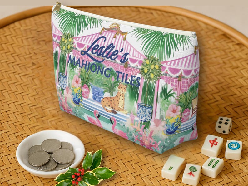 A Mahjong tile tote bag on a rattan surface beside Mahjong tiles, coins, and a sprig of holly. The design features pink, green, and blue tropical Chinoiserie design.