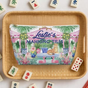 A personalized name Mahjong tile storage bag rests inside a bamboo tray surrounded by Mahjong tiles. The preppy chinoiserie design shows flamingos and a cheetah among tropical plants.