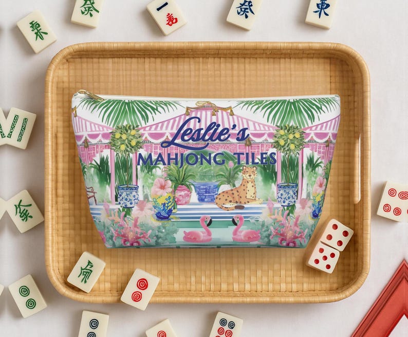 A personalized name Mahjong tile storage bag rests inside a bamboo tray surrounded by Mahjong tiles. The preppy chinoiserie design shows flamingos and a cheetah among tropical plants.