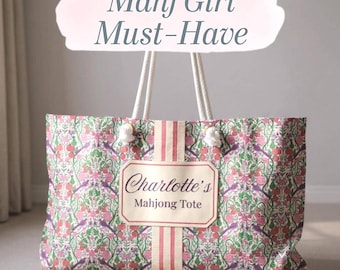 May include: A rectangular tote bag featuring a floral and bird pattern in shades of pink, green, and purple. It has white rope handles and a central cream-colored label with the text "Charlotte's Mahjong Tote". Above the bag, the text "Mahj Girl Must-Have" appears on a light pink brushstroke background.