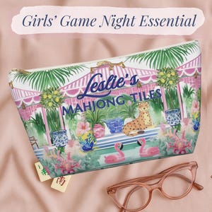 A colorful zip pouch printed with flamingos, tropical plants, and a cheetah under a pink cabana. The text “Leslie’s Mahjong Tiles” appears on the front. The bag rests on a pink cloth beside glasses and Mahjong tiles.