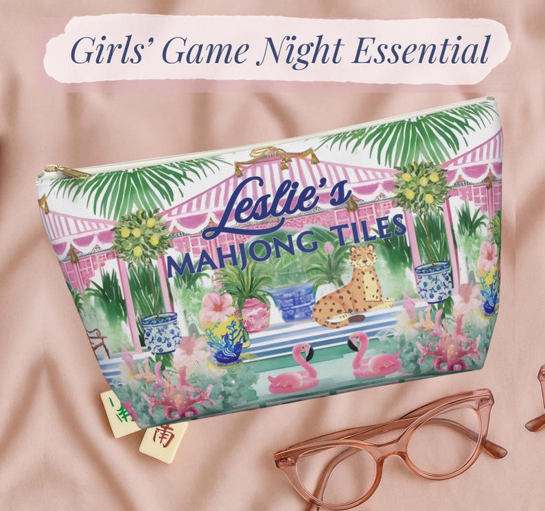 A colorful zip pouch printed with flamingos, tropical plants, and a cheetah under a pink cabana. The text “Leslie’s Mahjong Tiles” appears on the front. The bag rests on a pink cloth beside glasses and Mahjong tiles.