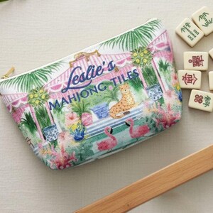 A chinoiserie, preppy Palm beach-style tropical print Mahjong tile storage pouch resting on a linen surface with Mahjong tiles nearby. The text “Leslie’s Mahjong Tiles” appears across the front to show personalization option