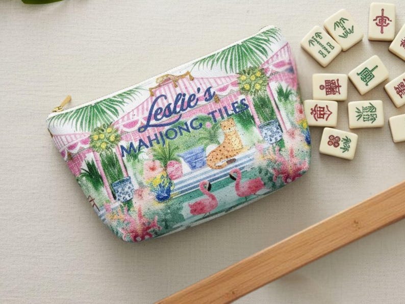 A chinoiserie, preppy Palm beach-style tropical print Mahjong tile storage pouch resting on a linen surface with Mahjong tiles nearby. The text “Leslie’s Mahjong Tiles” appears across the front to show personalization option