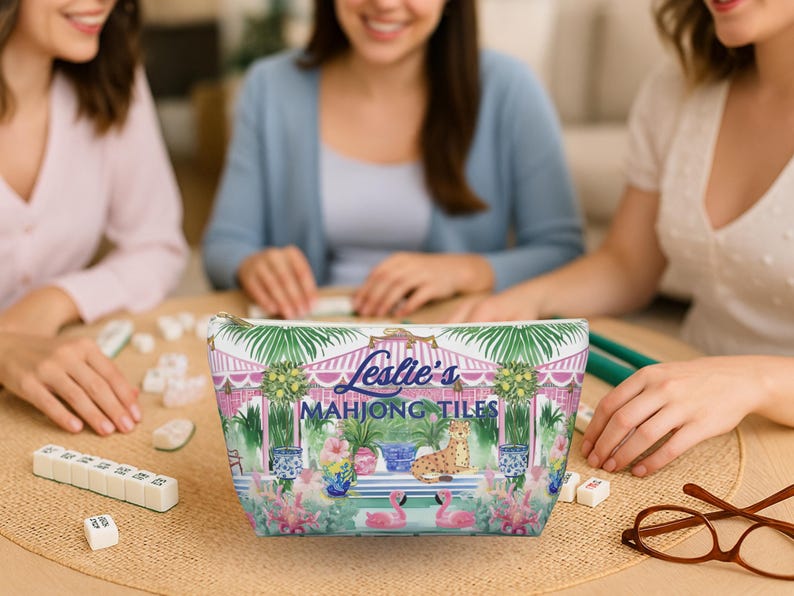 Three women sit around a table playing Mahjong. A colorful personalized Mahjong tile bag sits upright in the center of the table. Tile bag features a custom name and a preppy chinoiserie print with striped cabana, cheetah and potted lemon trees.