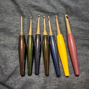 May include: A collection of seven crochet hooks with metallic silver tips and colorful handles. The handles are in shades of brown, green, blue, yellow, and purple. The hooks are arranged on a denim surface.