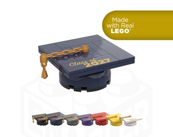May include: A dark blue graduation cap constructed from LEGO bricks, featuring a gold chain tassel and the text Class of 2027 printed in gold on the top surface. A row of seven additional graduation caps in various colors is displayed below.