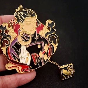 May include: Gold-tone enamel pin featuring a stylized portrait of a person with black hair and a red and black outfit. The pin has a chain attached to a small gold-tone charm with a stylized mouse design.