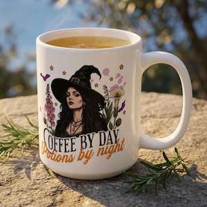 May include: A white ceramic mug featuring a graphic of a person wearing a black witch hat, surrounded by flowers and small stars. The text "COFFEE BY DAY Potions by night" is printed in black and orange lettering on the front of the mug.