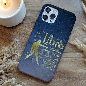 May include: A navy blue phone case with a Libra zodiac design. The case features a gold illustration of the Libra symbol and the word "Libra" in gold lettering, along with descriptive words. The case is on a wooden surface.