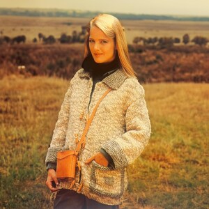 May include: A woman wearing a beige and gray knit jacket with a dark collar and trim. The jacket has large pockets and wooden toggle closures. A small, tan leather crossbody bag hangs from her shoulder. The background is a field with a muted color palette.