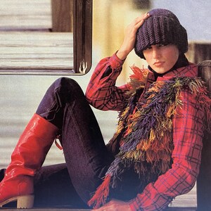 May include: A woman wearing a red and black plaid shirt with a colorful fringed vest, blue jeans, and red boots. She is wearing a dark purple knit hat.