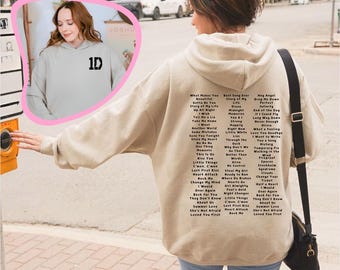 May include: A beige hoodie with a black print of a song list on the back. The list includes song titles such as "What Makes You Beautiful", "Best Song Ever", "My Angel", "Drag Me Down", "Perfect", "Up All Night", "Memories", "If I Could Fly", "Tell Me a Lie", "Long Way Down", "Take Me Home", "Strong", "Another World", "Little White Lies", "Love You Goodbye", "Save You Tonight", "Why Don't We", "Moments", "Temporary Pig", "Steal My Girl", "Ready to Run", "Where Do Broken Hearts Go", "Girl Almighty", "Fool's Gold", "Might Changes", "Little Things", "Last First Kiss", "Heart Attack", "Rock Me", "Change My Mind", "I Would", "Over Again", "Back for You", "They Don't Know About Us", "Summer Love", "She's Not Afraid", "Loved You First".