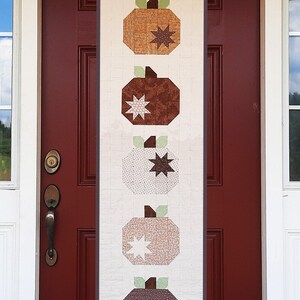 Puede incluir: Una puerta blanca con una puerta roja en el interior. La puerta roja tiene un panel largo y estrecho con un diseño de cinco calabazas marrones, blancas y verdes con estrellas.