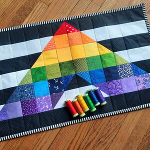 May include: A handmade quilt with a rainbow triangle design, set against a black and white striped background. The quilt features a variety of colorful squares and is bordered with a black and white striped pattern. Several spools of thread in matching colors are placed near the quilt.