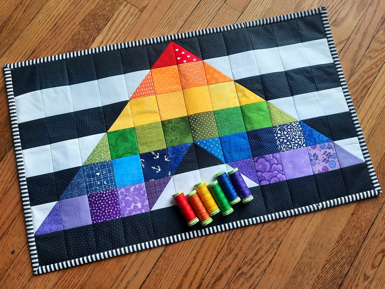 May include: A handmade quilt with a rainbow triangle design, set against a black and white striped background. The quilt features a variety of colorful squares and is bordered with a black and white striped pattern. Several spools of thread in matching colors are placed near the quilt.
