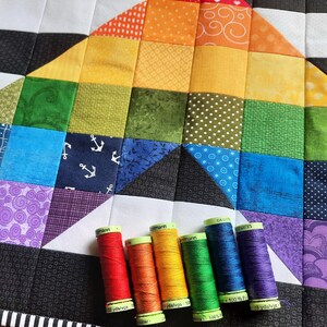 May include: A colorful quilt with a rainbow design, featuring squares of various patterns and colors. Six spools of thread in matching rainbow hues are arranged at the bottom. The thread spools have text that reads "G&uuml;termann".