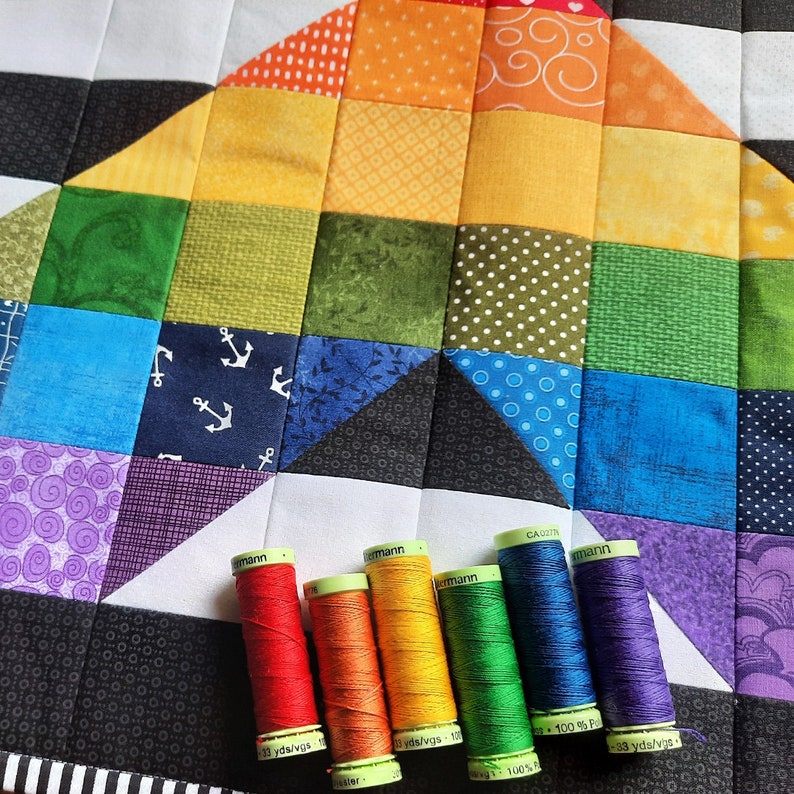 May include: A colorful quilt with a rainbow design, featuring squares of various patterns and colors. Six spools of thread in matching rainbow hues are arranged at the bottom. The thread spools have text that reads "G&uuml;termann".