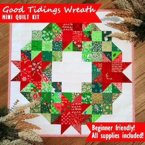 May include: A square mini quilt kit featuring a Christmas wreath design. The wreath is composed of red and green fabric squares with festive patterns. The text "Good Tidings Wreath" and "Mini Quilt Kit" are displayed at the top. The text "Beginner friendly! All supplies included!" is at the bottom.