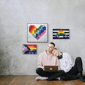 May include: Three colorful, framed wall hangings with rainbow designs, including a heart, a triangle, and a Pride flag. Two people are sitting on the floor, one using a laptop. The wall is gray, and the floor is wood.
