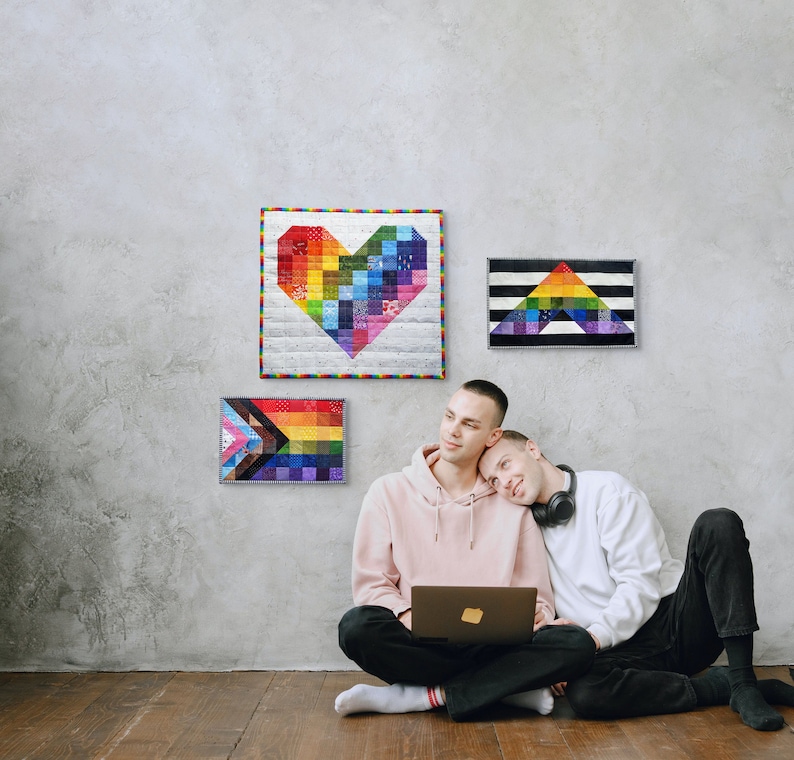 May include: Three colorful, framed wall hangings with rainbow designs, including a heart, a triangle, and a Pride flag. Two people are sitting on the floor, one using a laptop. The wall is gray, and the floor is wood.