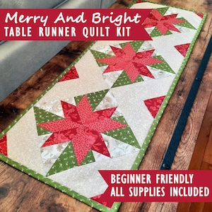 Op de afbeelding: Een rechthoekige tafelloper quiltkit met een herhalend sterpatroon in rode en groene stof met crèmekleurige accenten. De tekst Merry And Bright TABLE RUNNER QUILT KIT en BEGINNER FRIENDLY ALL SUPPLIES INCLUDED is zichtbaar.