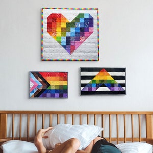 May include: Three colorful, quilted wall hangings in a bedroom. The top one is a rainbow heart shape. The other two are rectangular, one with a rainbow flag design and the other with a rainbow triangle design. The bed has white pillows and two people.