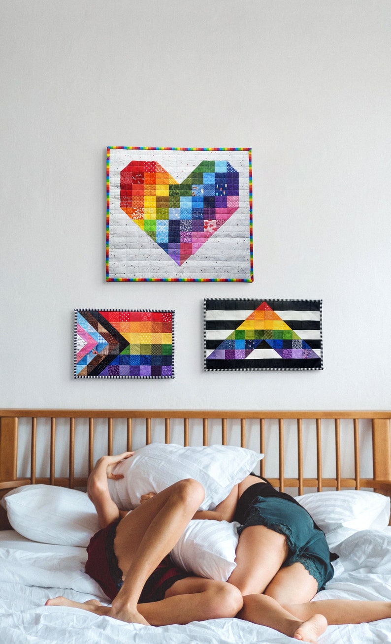 May include: Three colorful, quilted wall hangings in a bedroom. The top one is a rainbow heart shape. The other two are rectangular, one with a rainbow flag design and the other with a rainbow triangle design. The bed has white pillows and two people.