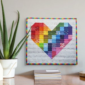 May include: A rainbow heart-shaped quilt with a white background and a black and white speckled pattern. The quilt is framed with a rainbow border.