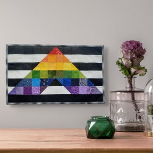 May include: A colorful, handmade quilt featuring a rainbow triangle design. The triangle is set against a black and white striped background. The quilt is displayed on a wall, with decorative items on a wooden surface in the foreground.