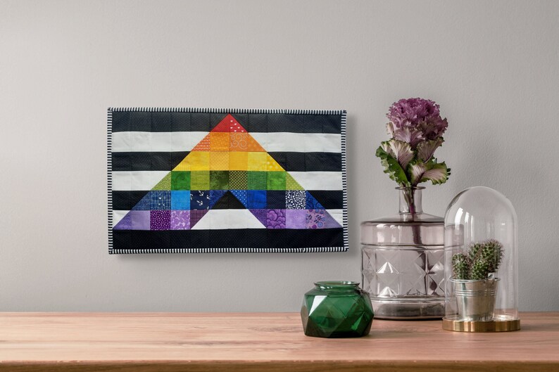 May include: A colorful, handmade quilt featuring a rainbow triangle design. The triangle is set against a black and white striped background. The quilt is displayed on a wall, with decorative items on a wooden surface in the foreground.