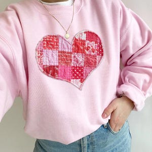 May include: A pink sweatshirt with a large patchwork heart design. The heart is made of pink and red fabric squares with the word "love" printed on some of the squares.