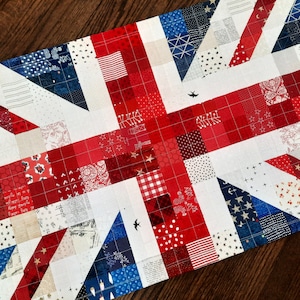 May include: A quilted table runner featuring a patchwork design in red, white, and blue, resembling the Union Jack flag. The runner is made of various patterned fabrics, including stars, stripes, and polka dots. Two small bird silhouettes are stitched on the white background.